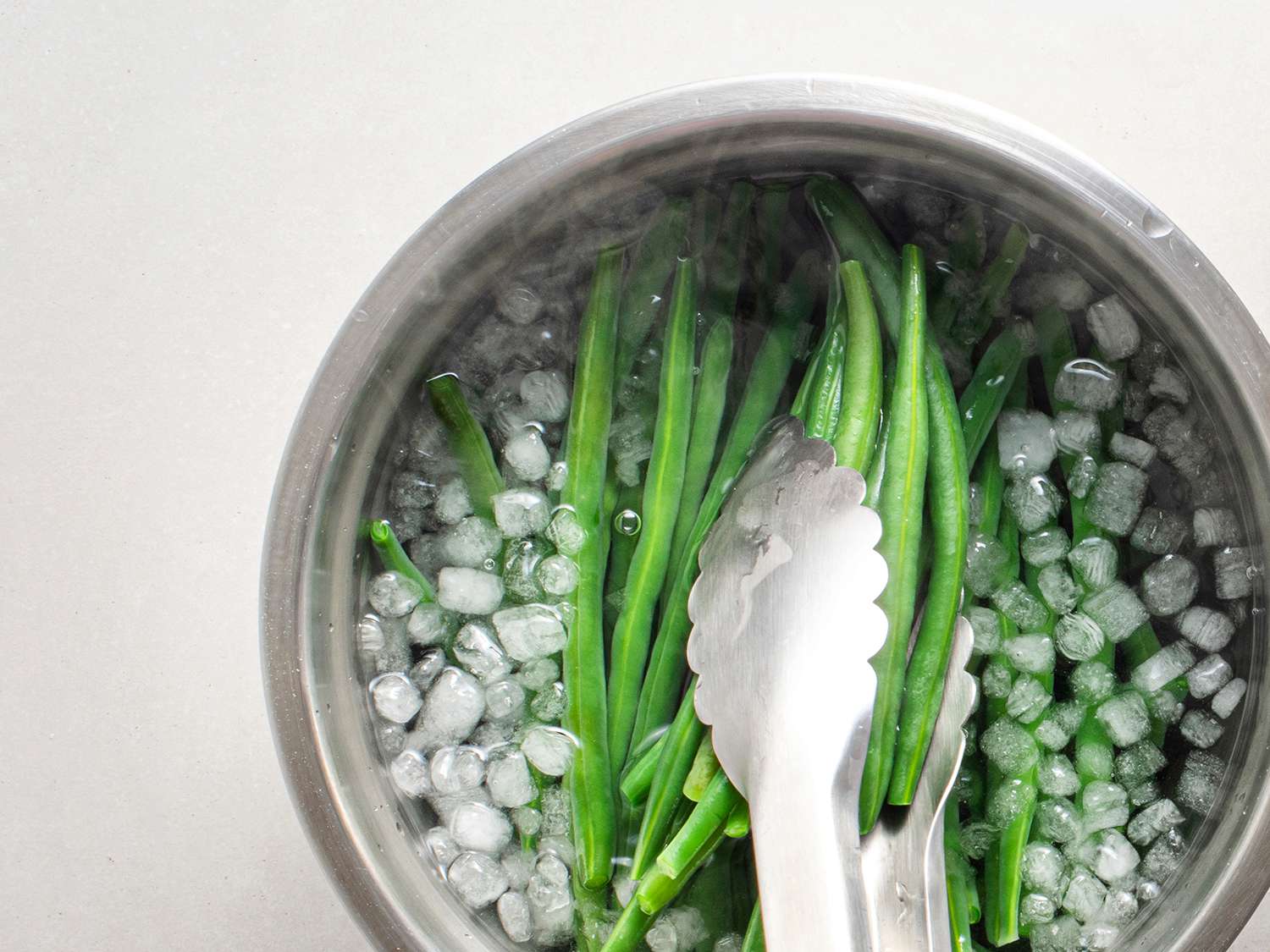 Blanched green beans being chilled in an ice bath, and lifted out with a pair of tongs.