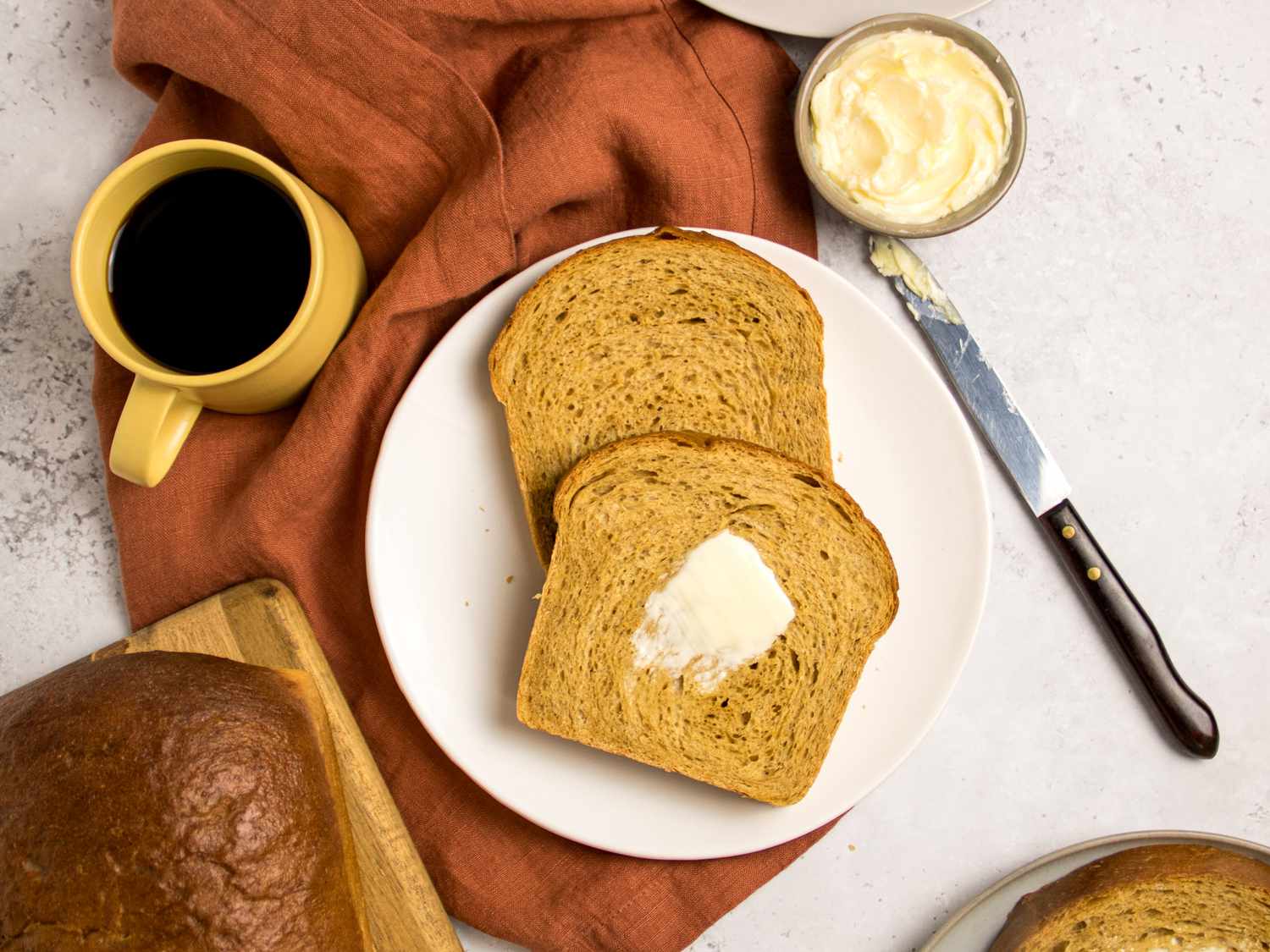 Sliced loaf of anadama bread on a wooden board, with two slices on plates, with rust colored napkin, mug of coffee, plate, butter, and breadknife behind it 