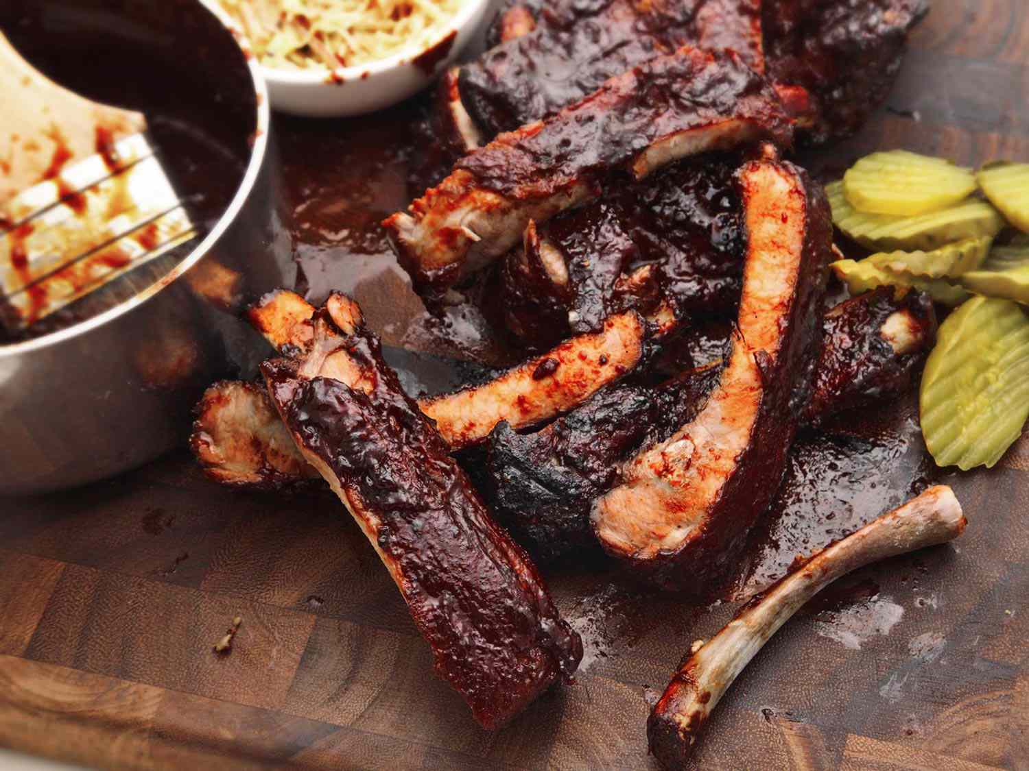 Pork ribs cooked sous vide, coated in barbecue sauce, next to pickle slices on a wooden board.