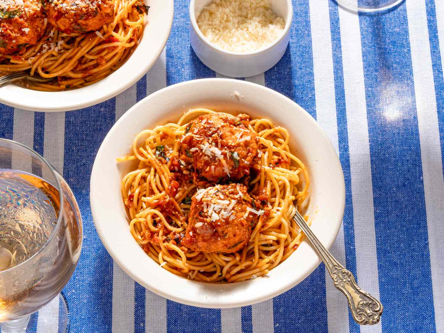 A bowl of spaghetti with chicken meatballs and grated Parmesan on a table with a blue striped tablecloth