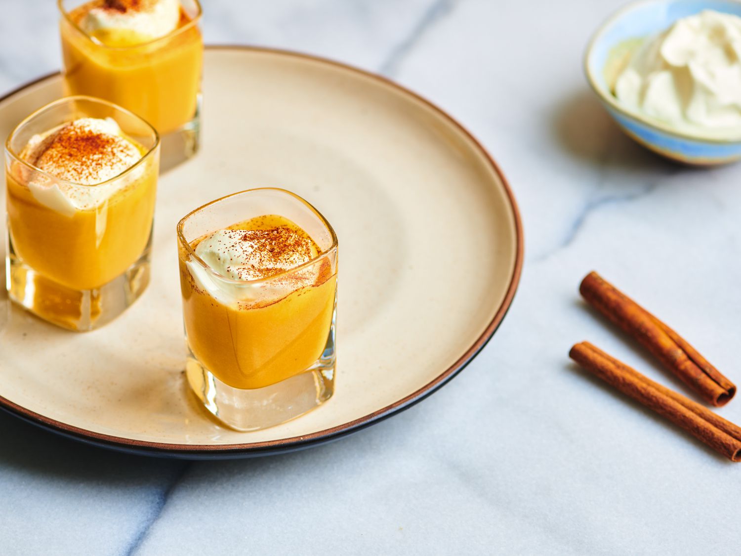 Three small glasses filled with DIY Advocaat and topped with whipped cream and dusted with cinnamon, on a ceramic plate. On the right periphery are a couple of sticks of cinnamon and a small bowl of whipped cream.