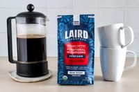 A French press, a package of Laird Superfood coffee, and stacked white mugs on a countertop