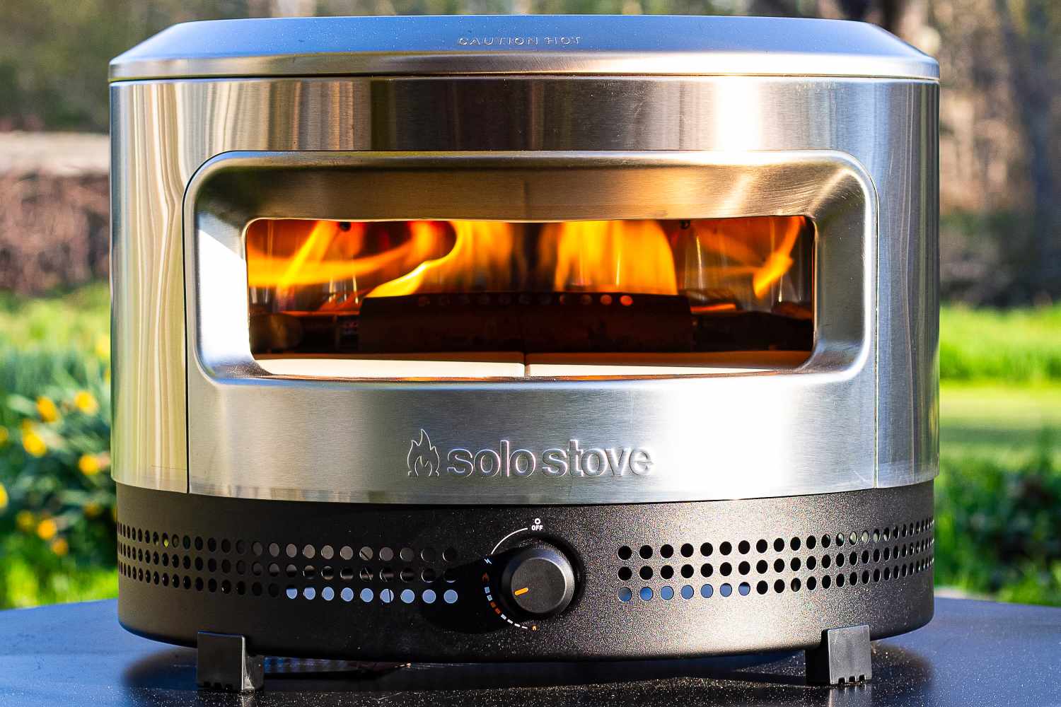 A Solo Stove Pi Prime with visible flames inside, standing on a surface with a blurred garden background