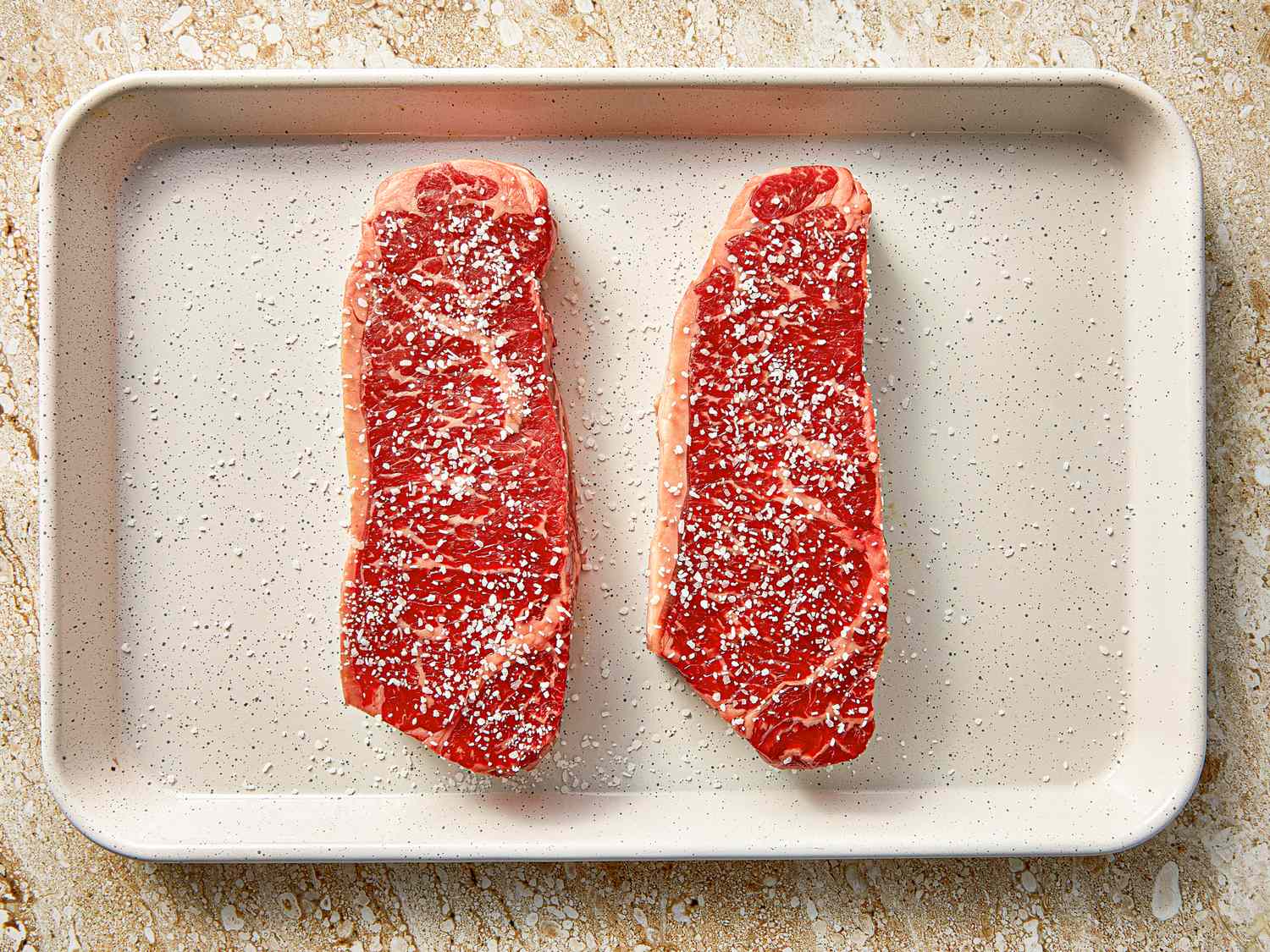 Two raw steaks seasoned with salt on a baking tray