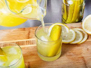 Glass of lemonade with a pickle spear lemon slices and a mason jar with pickles nearby on a wooden surface