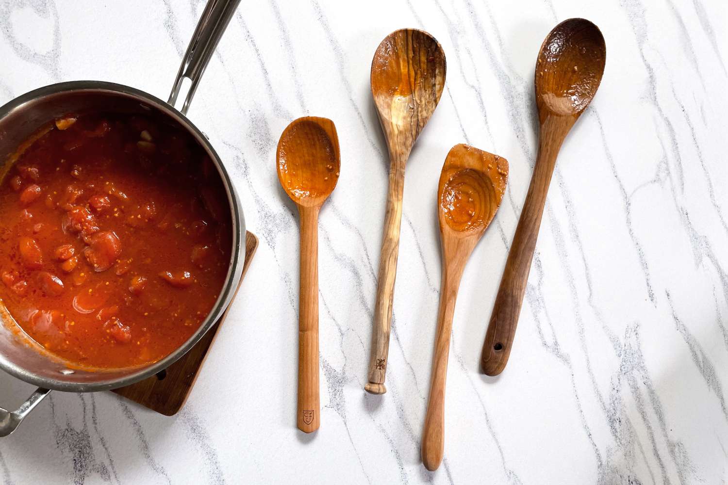 A pot of tomatobased sauce and four different wooden spoons on a marble countertop