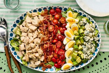 A Cobb salad served on a decorative platter, featuring rows of ingredients including chicken, bacon, cherry tomatoes, eggs, avocado, and blue cheese