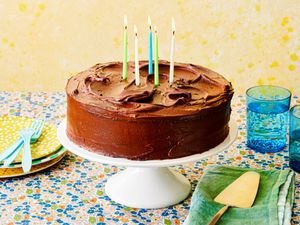 A chocolate-frosted layer cake with billowing swirls of frosting, birthday candles, accompanied by serving plates, forks, and blue glasses of water against a yellow background.