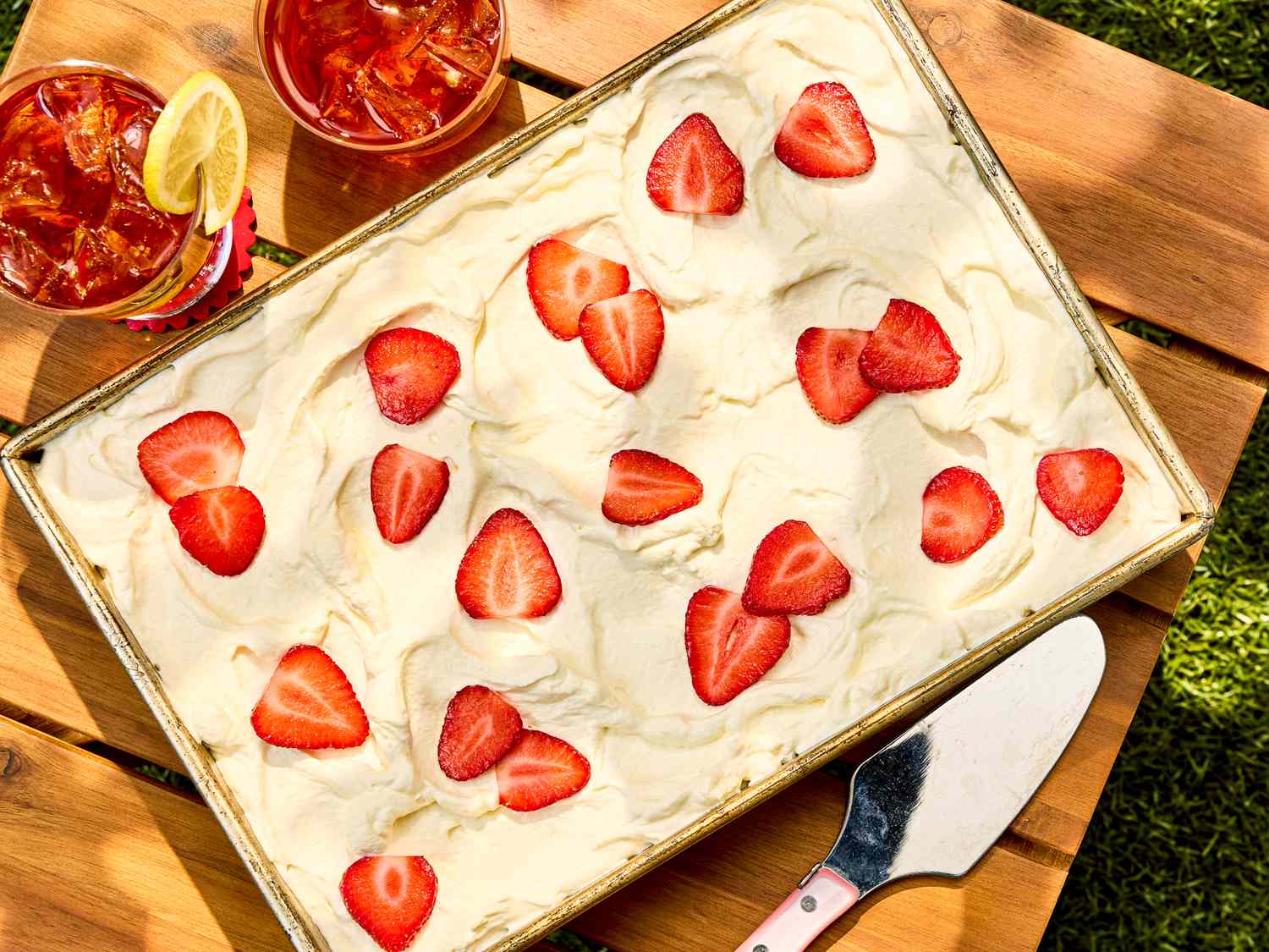 A dessert topped with strawberries on a wooden table accompanied by drinks and a serving spatula