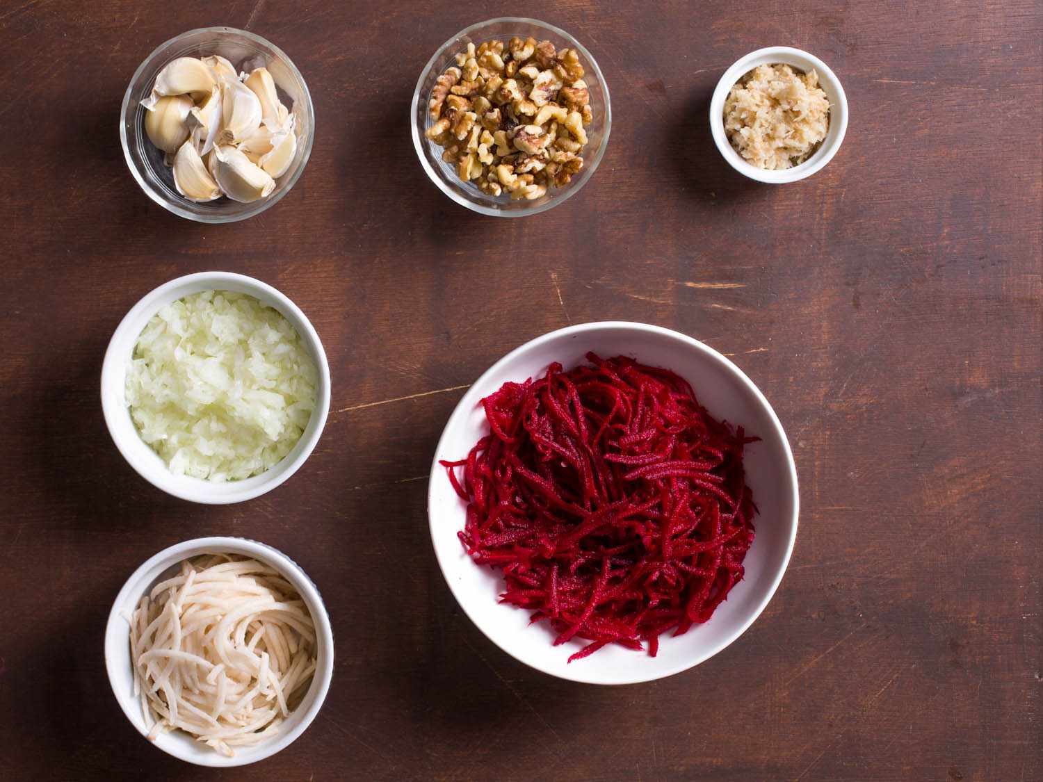 Overhead shot of ingredients for beet-horseradish latkes: shredded beet, shredded potato, chopped onion, garlic cloves, walnuts, and horseradish