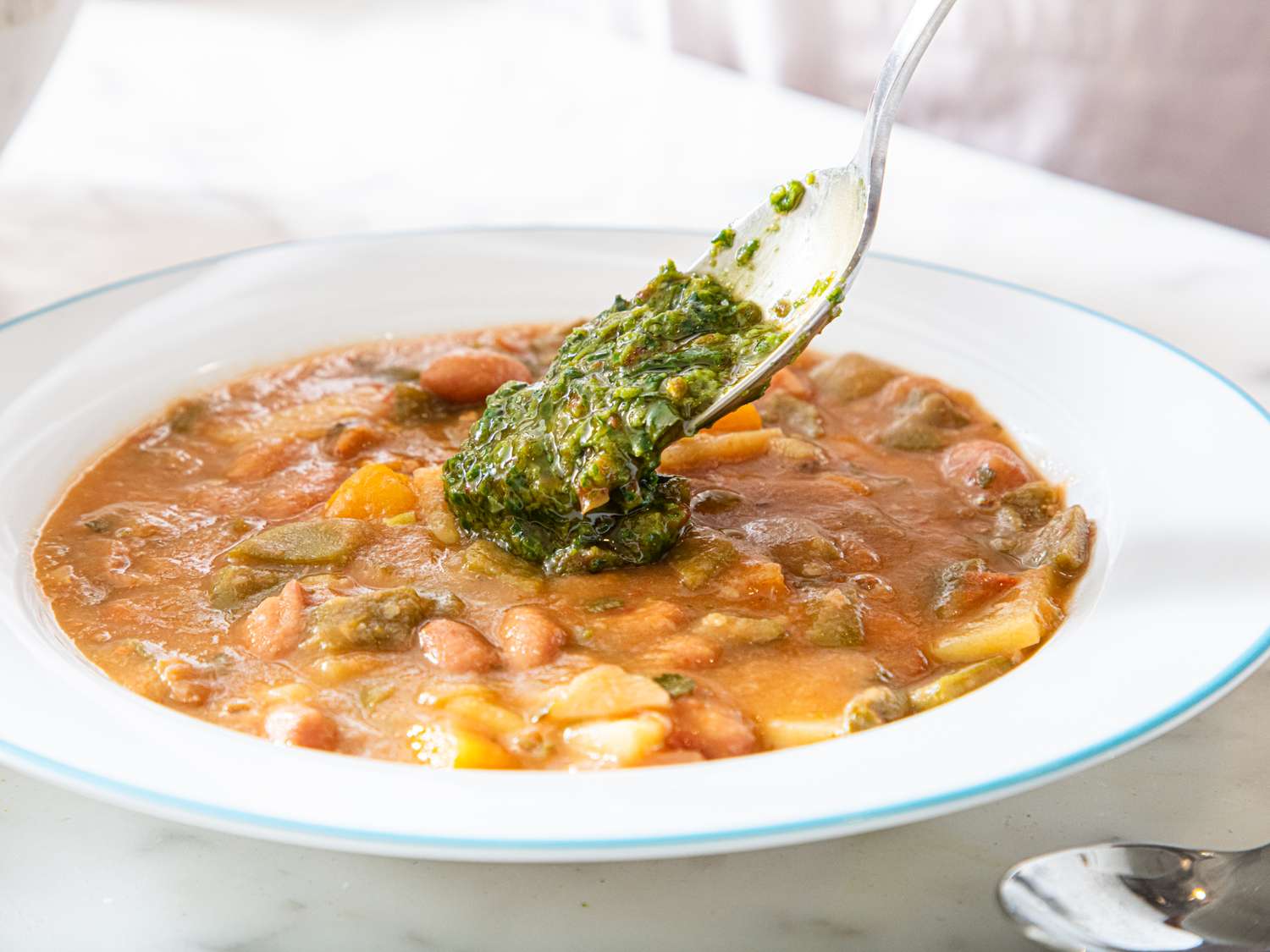 A bowl of vegetable soup being garnished with a green herb mixture a spoon adding the topping