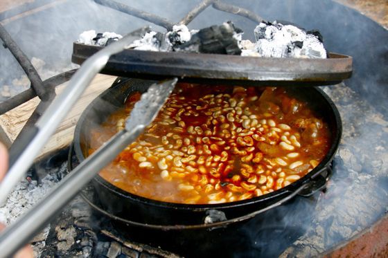 Using tongs to peek under the lid of a Dutch oven filled with bubbling chili over campfire coals.