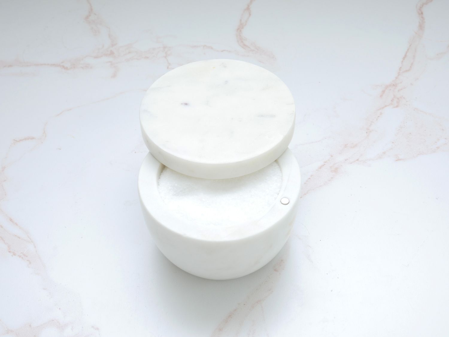 Marble salt cellar with lid slightly ajar, salt inside, on a pink and white marble countertop