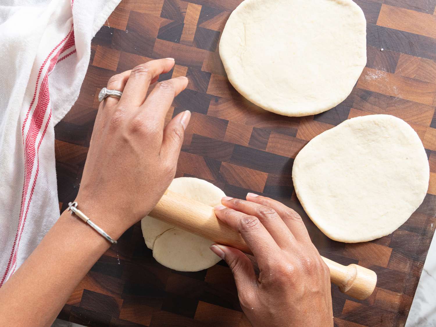Flattened Johnny Cake dough rolls on a wooden cutting board, with hands holding one cake and a wooden rolling pin 