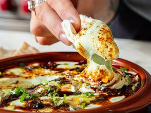 A hand dipping a piece of bread into a baked cheese and herb dish served in a terracotta bowl