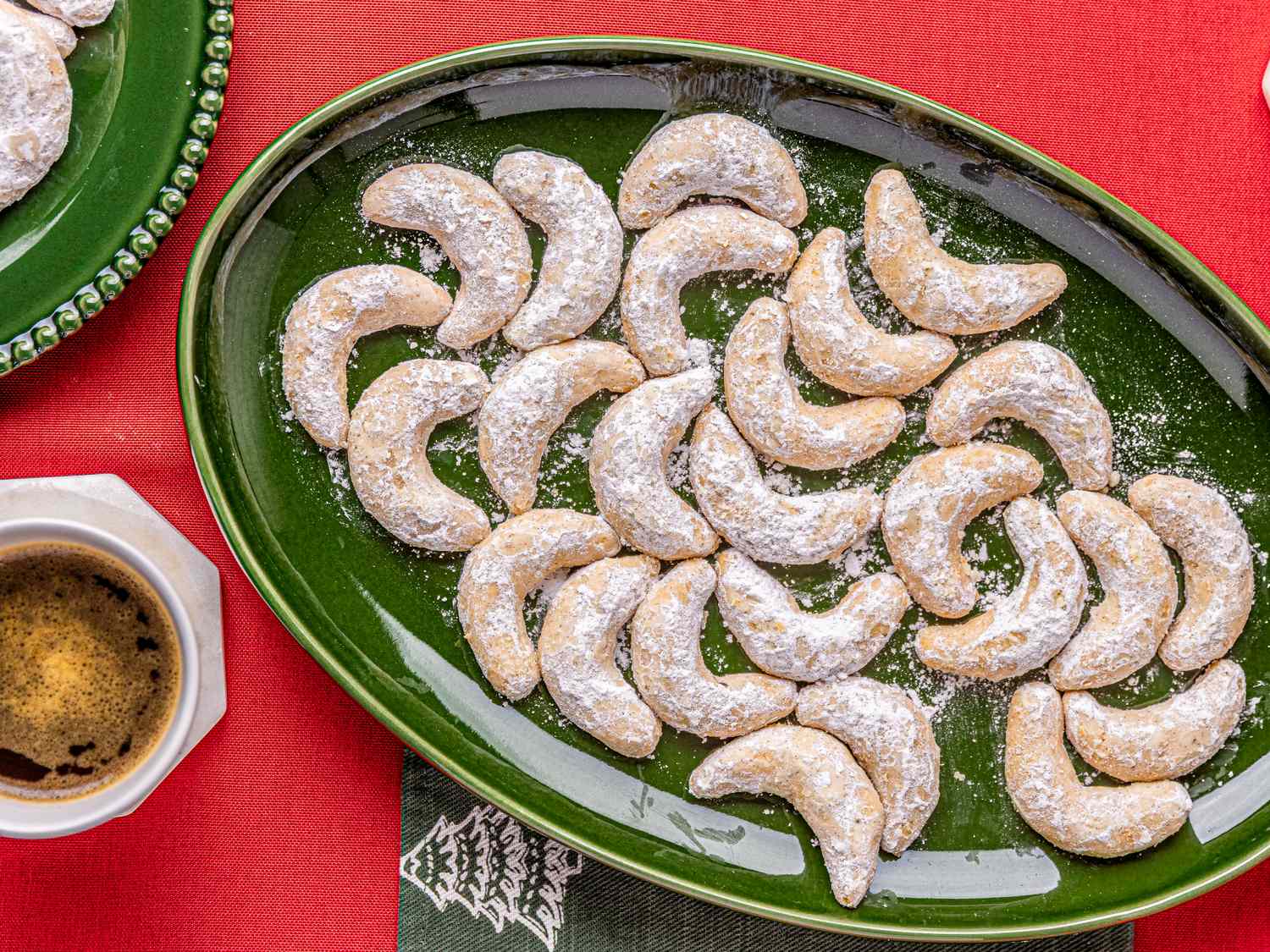 A plate of crescentshaped cookies covered in powdered sugar displayed on a green platter with a cup of coffee nearby