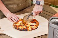 A person cutting a pizza made in the Ninja Woodfire 8-in-1 Outdoor Oven