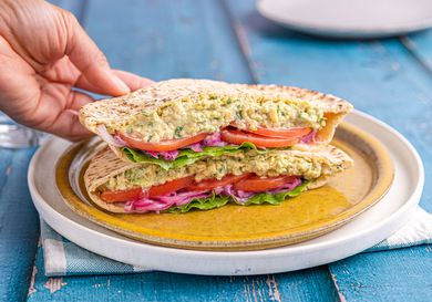 A hand holding a chickpea salad sandwich cut in half, filled with lettuce, red onion, and tomato, placed on a plate
