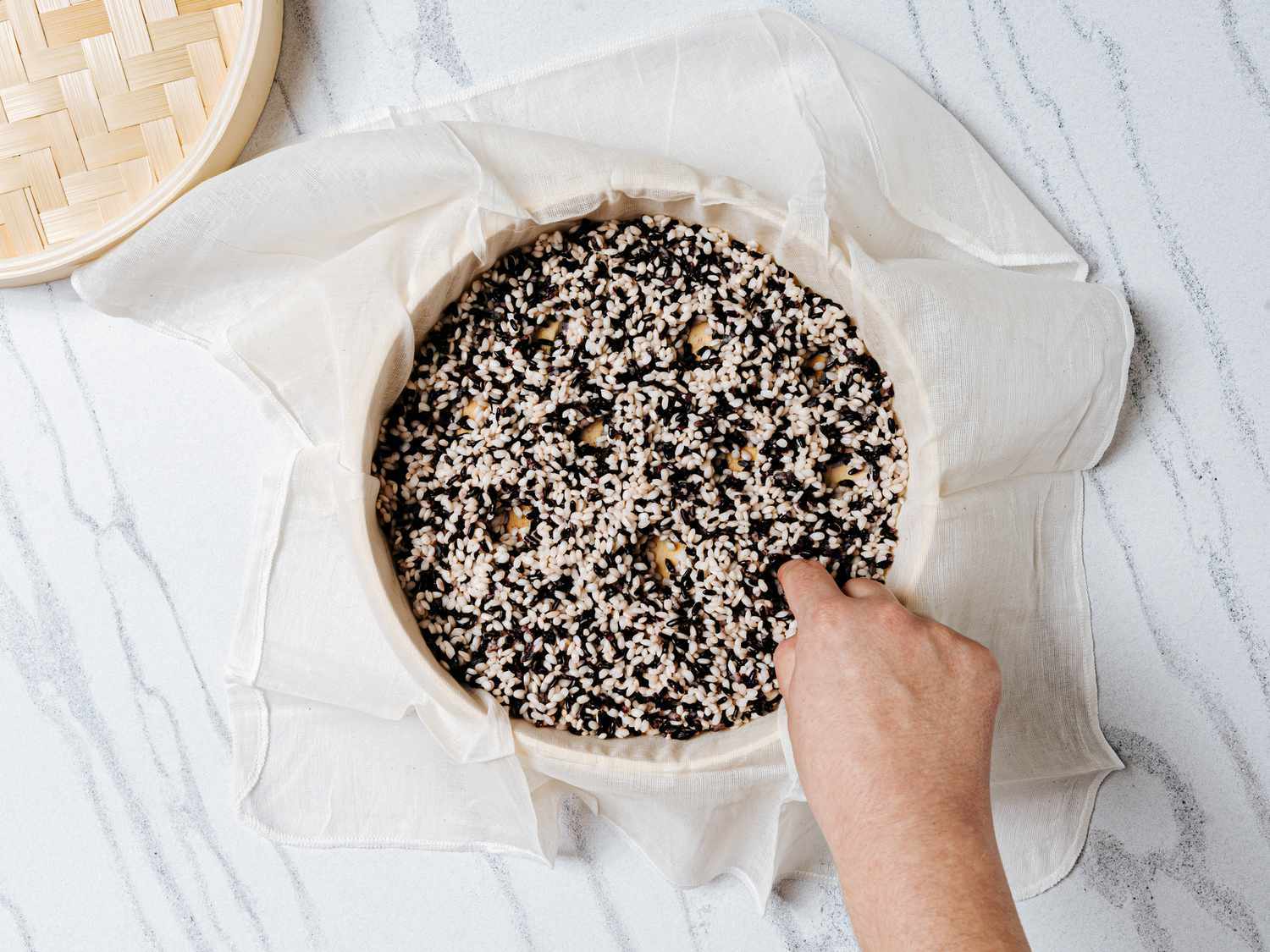 rice lined up on a cheesecloth inside a bamboo steamer. A hand flattening the rice evenly.