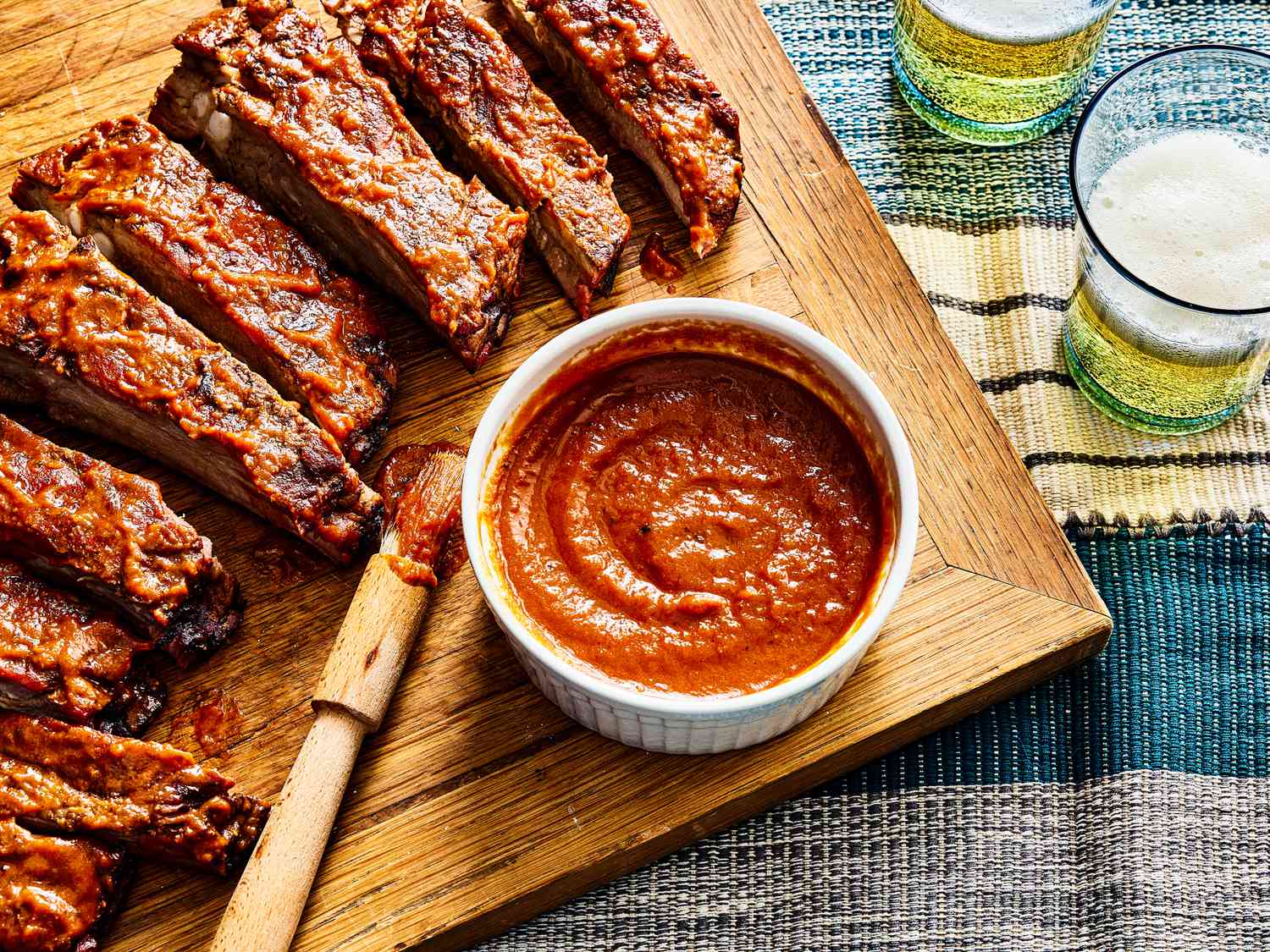 overhead view of bbq sauce on a cutting board alongside ribs and glasses of beer