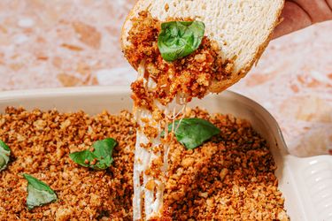 A bread slice dipped into a casserole with melted cheese and breadcrumbs garnished with fresh basil leaves