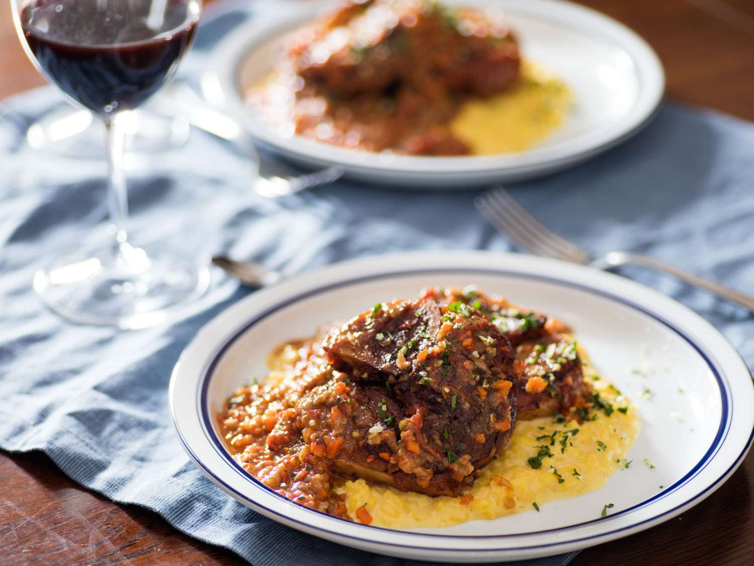 Plate of osso buco on top of risotto alla milanese with second serving and glass of wine in the background.