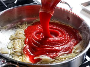 Tomato puree being poured into a pan with sautéed onions on a stovetop surface