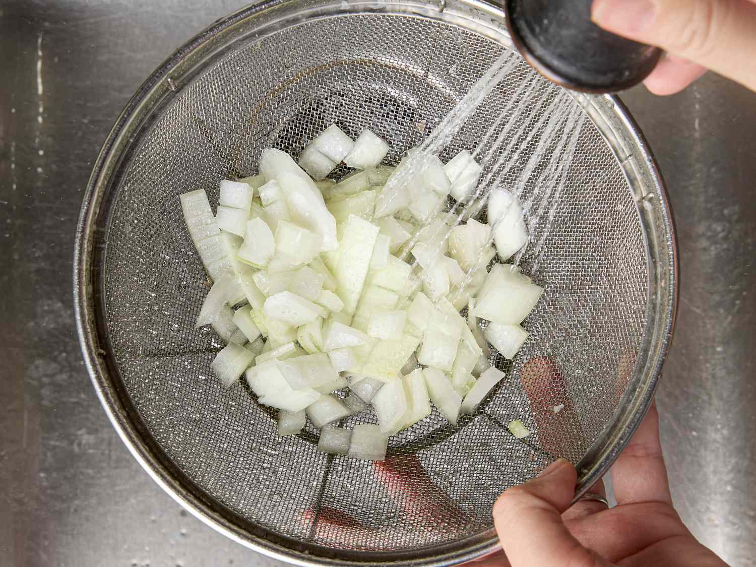 Diced onions being rinsed in a metal sieve under running water in a kitchen sink