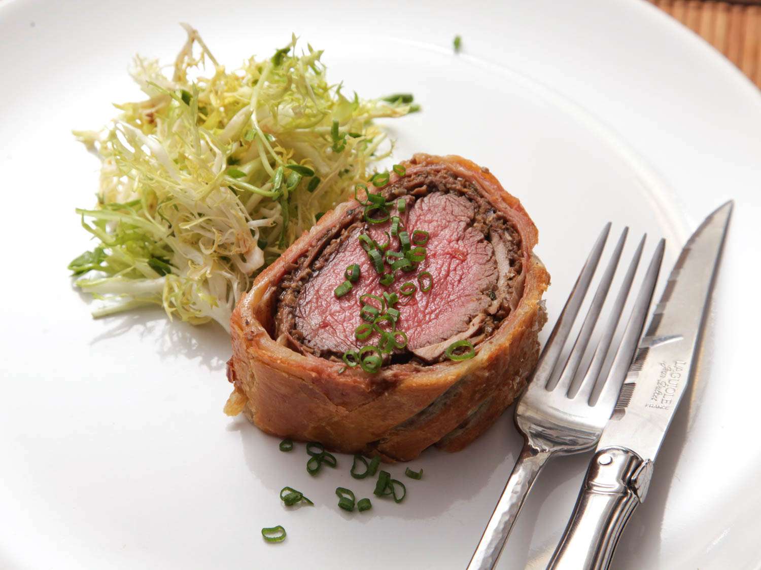 Closeup of a thick slab of beef Wellington, served on a porcelain plate with a tangle of dressed frisée.