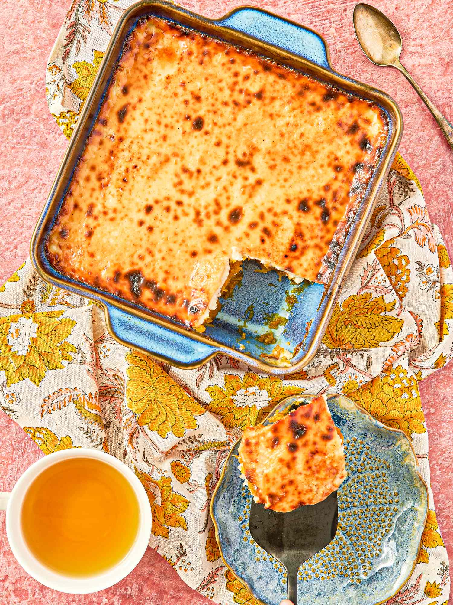 A baked dish with a portion served onto a blue plate accompanied by a cup of tea