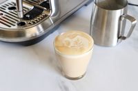 A latte with latte art in a glass next to an espresso machine and a metal frothing pitcher on a countertop