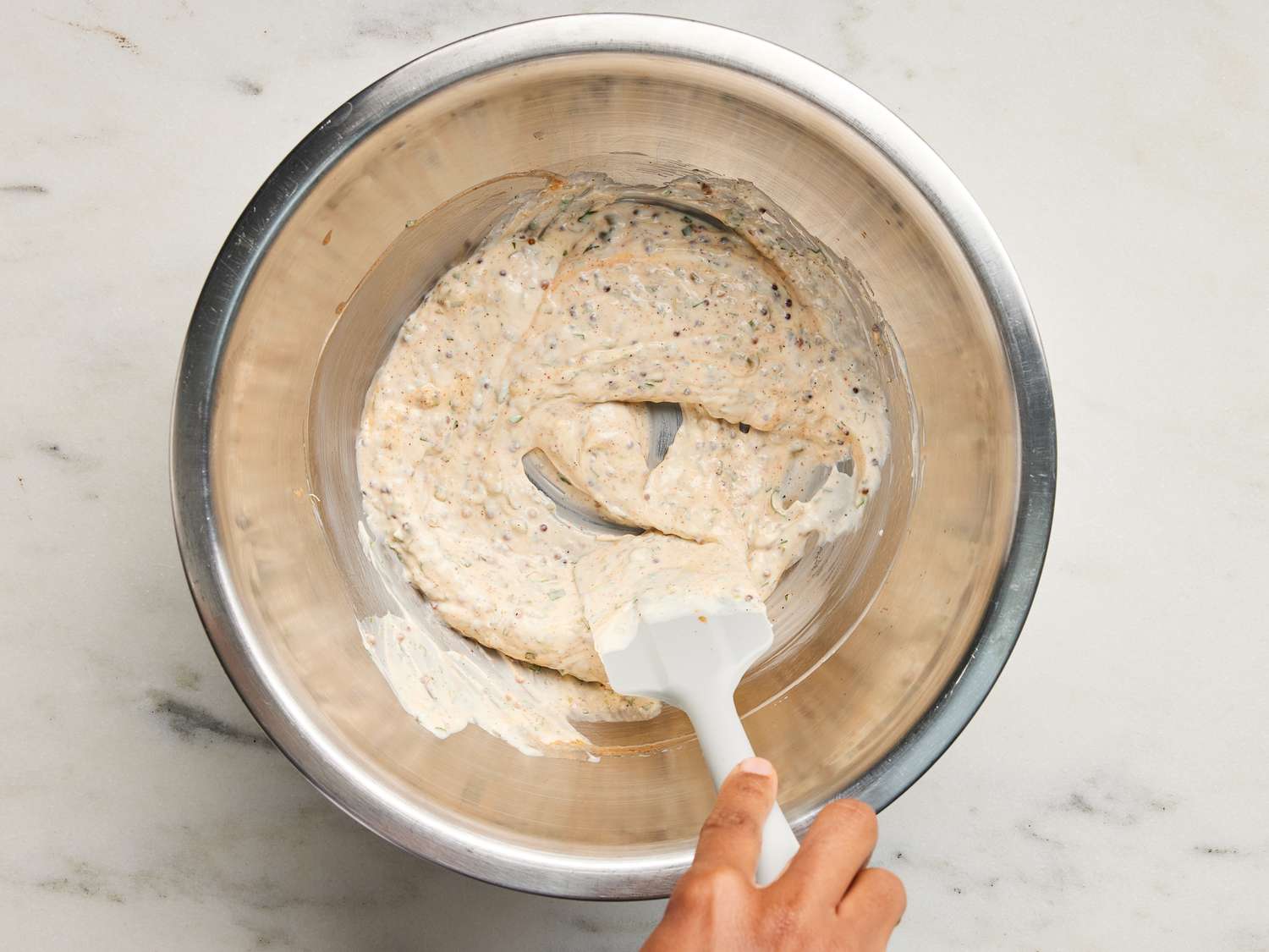 Rémoulade mixture being stirred with silicone spatula in a metal bowl on a marble surface 