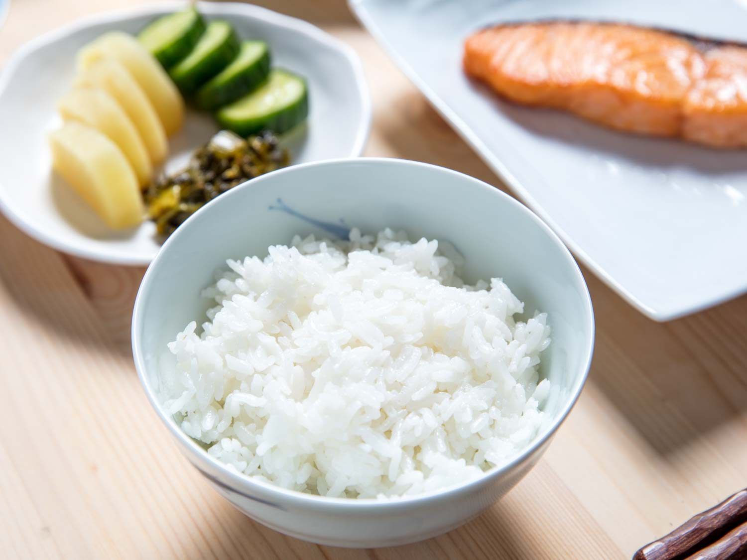 A ceramic bowl of white rice, with pickles and salted salmon in the background