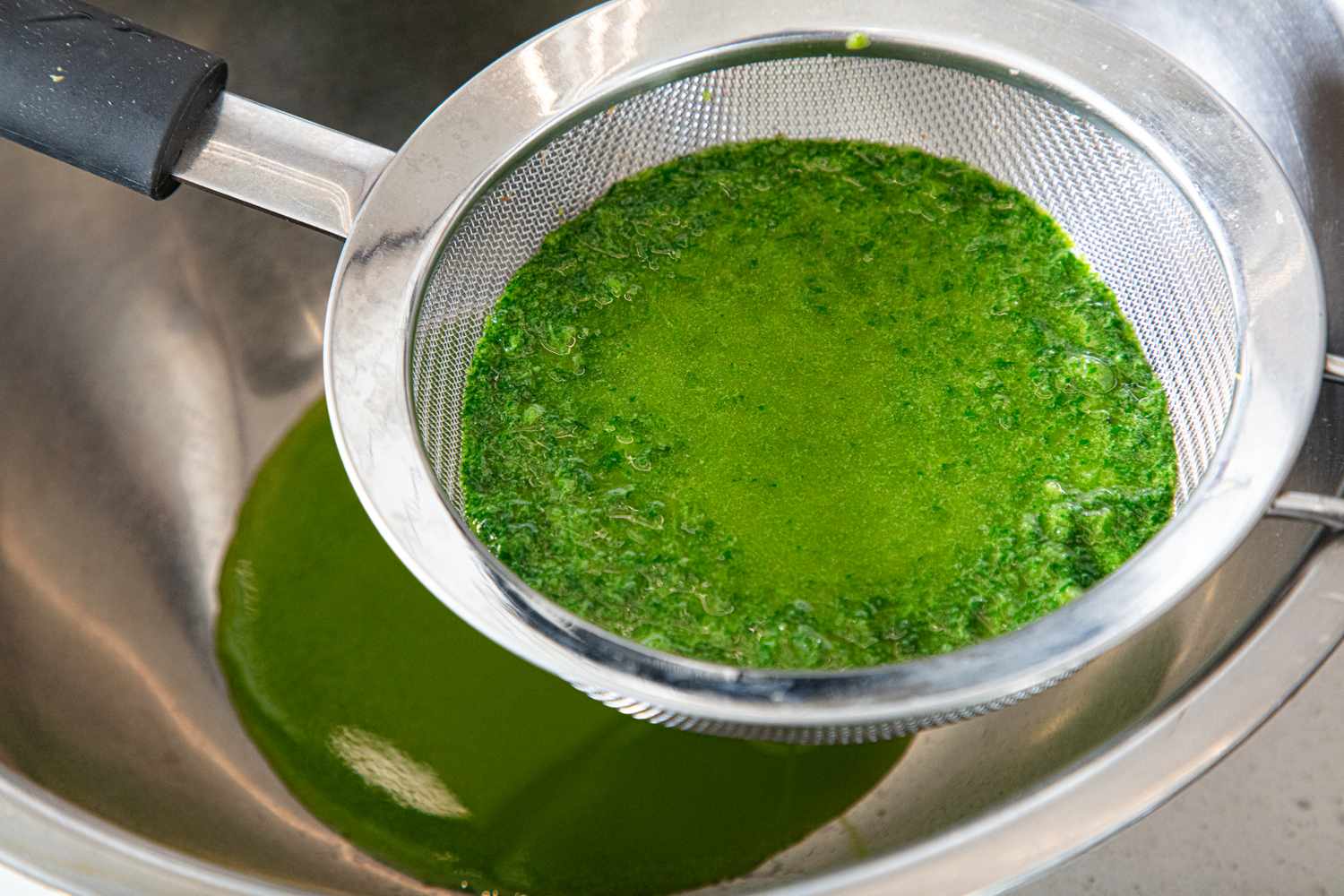 A metal strainer with blended green herbs over a bowl oil or liquid filtered below in the bowl