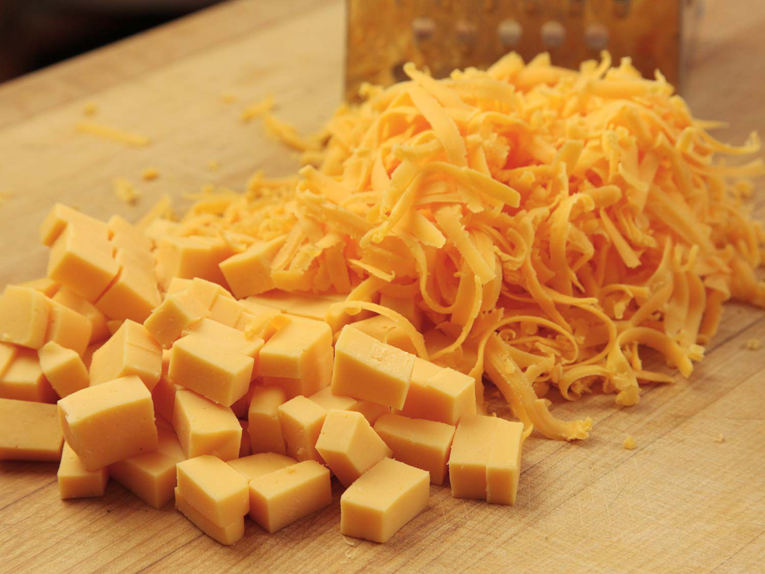 Cheddar cheese shredded with a box grater next to cubes of American cheese.