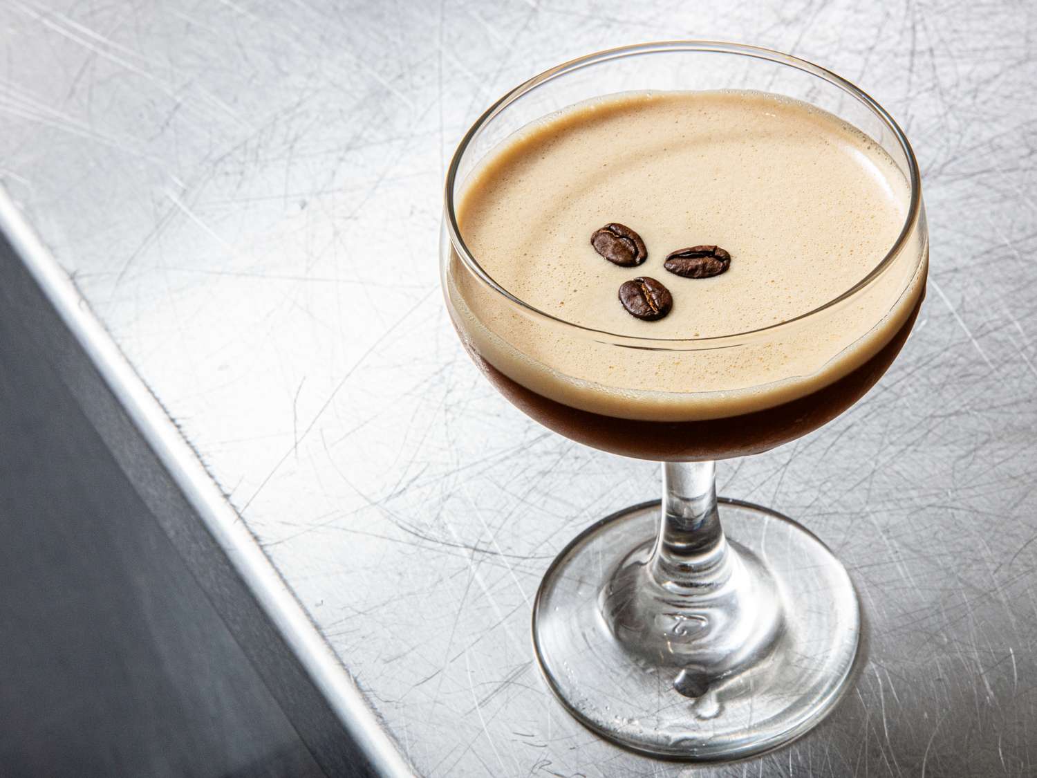 A glass of espresso martini garnished with three coffee beans on a metallic surface