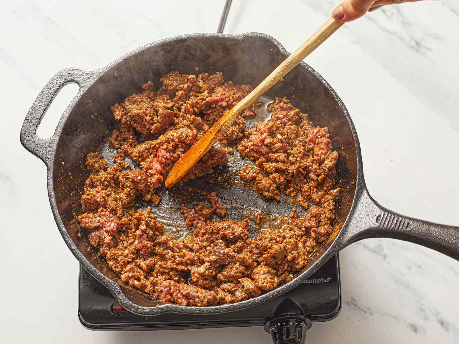 Ground meat cooking in a cast iron skillet, being stirred with a wooden spoon.