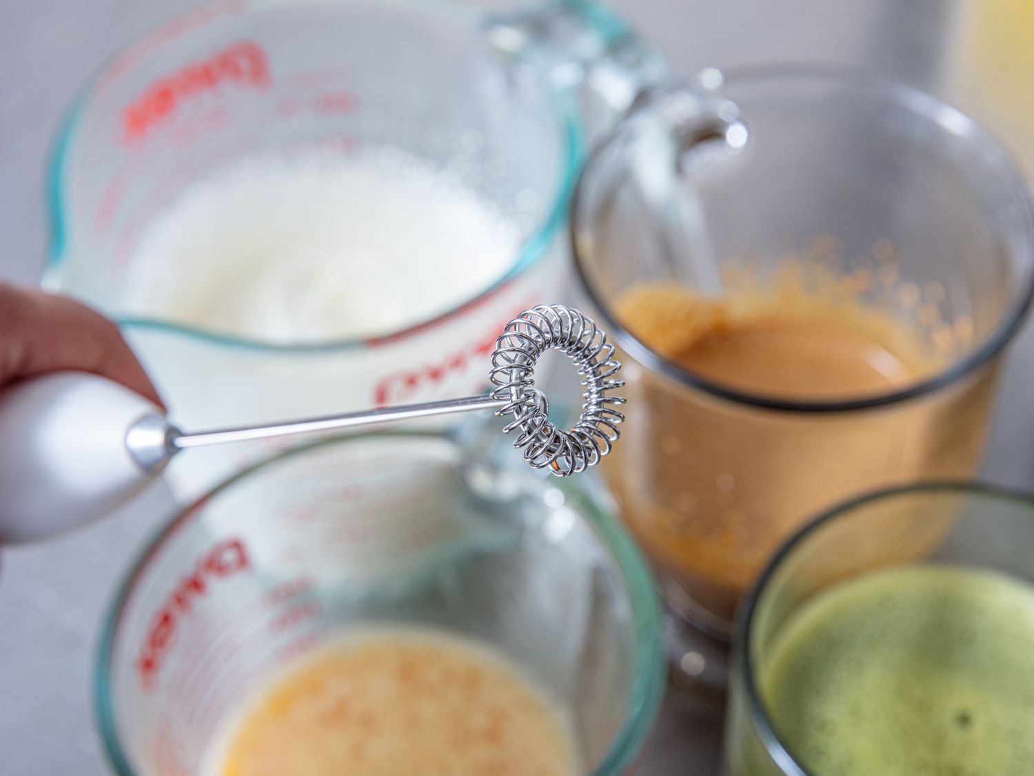 Hand using a milk frother above glasses containing beverages cups and prepared drinks visible