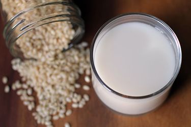 A glass of DIY rice milk. A glass canning jar of dry rice grains has been artfully spilled on the countertop nearby.