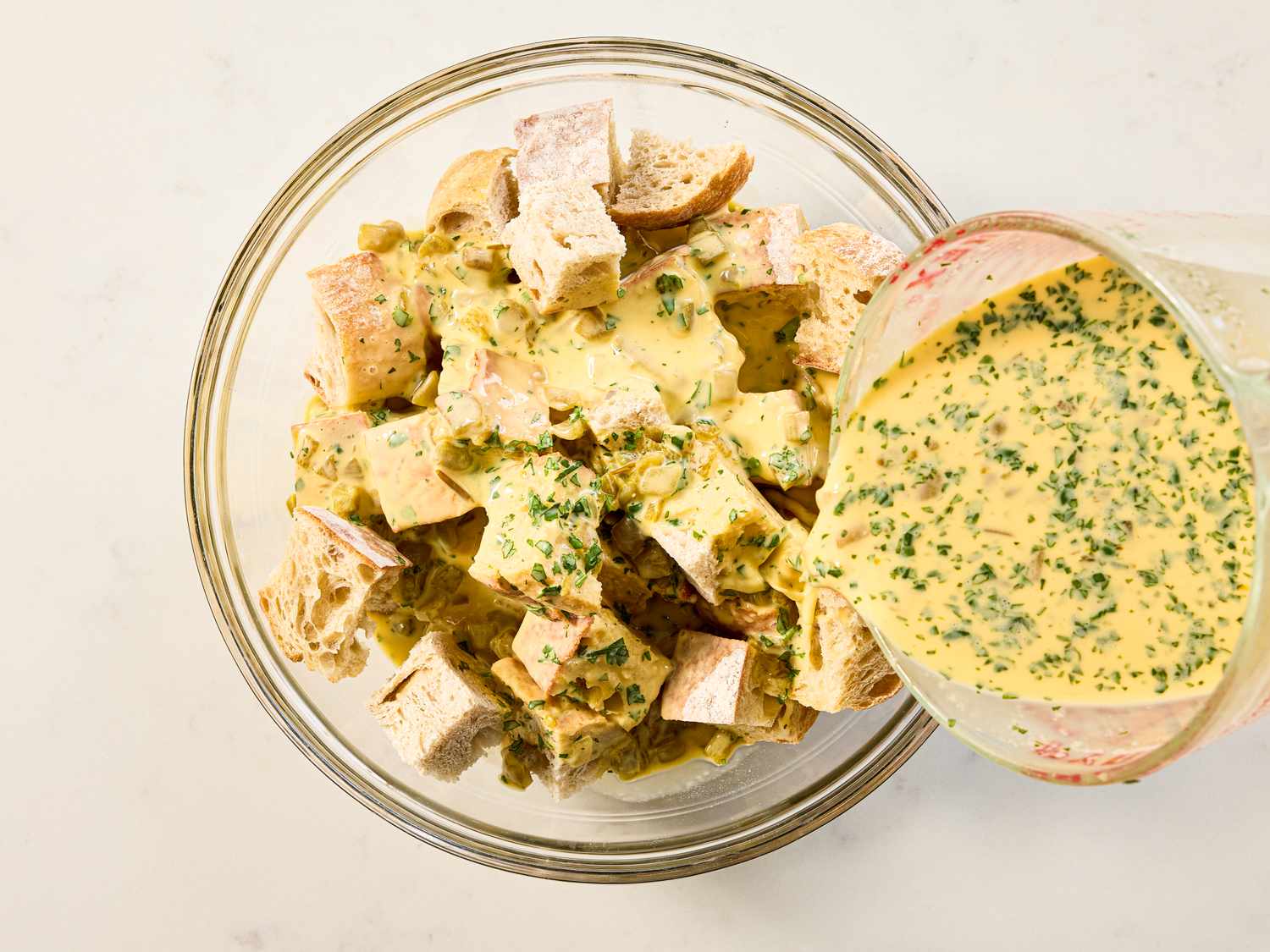 Preparation of a breakfast strata with bread cubes being mixed with a sauce containing spices and herbs in a glass bowl