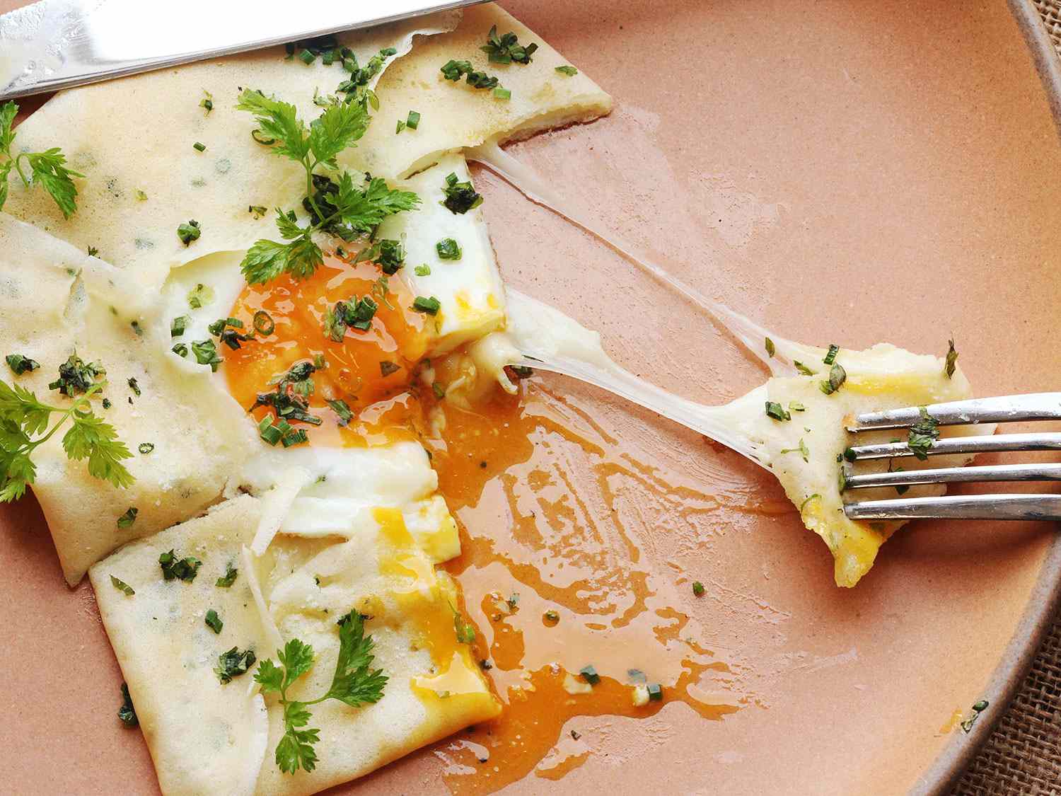 Close-up of a half-eaten French crepe with ham, cheese, and egg. A fork drags a cheesy bite across the plate.