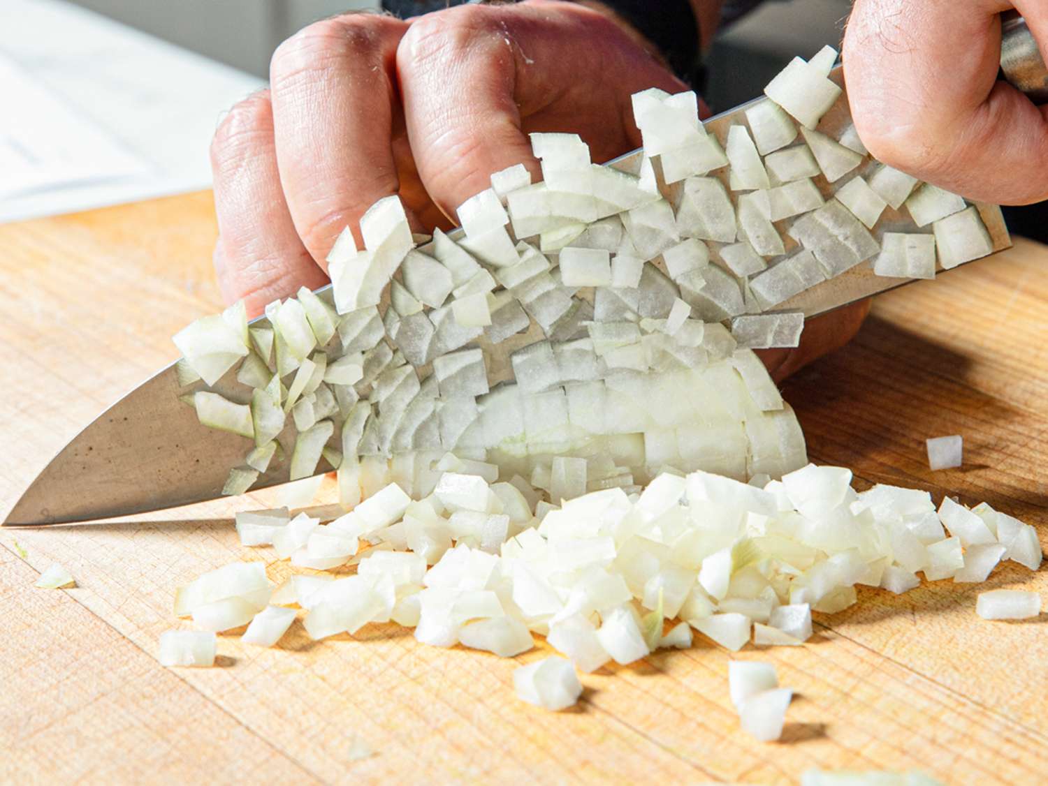 A person finely dicing an onion with a knife on a wooden cutting board