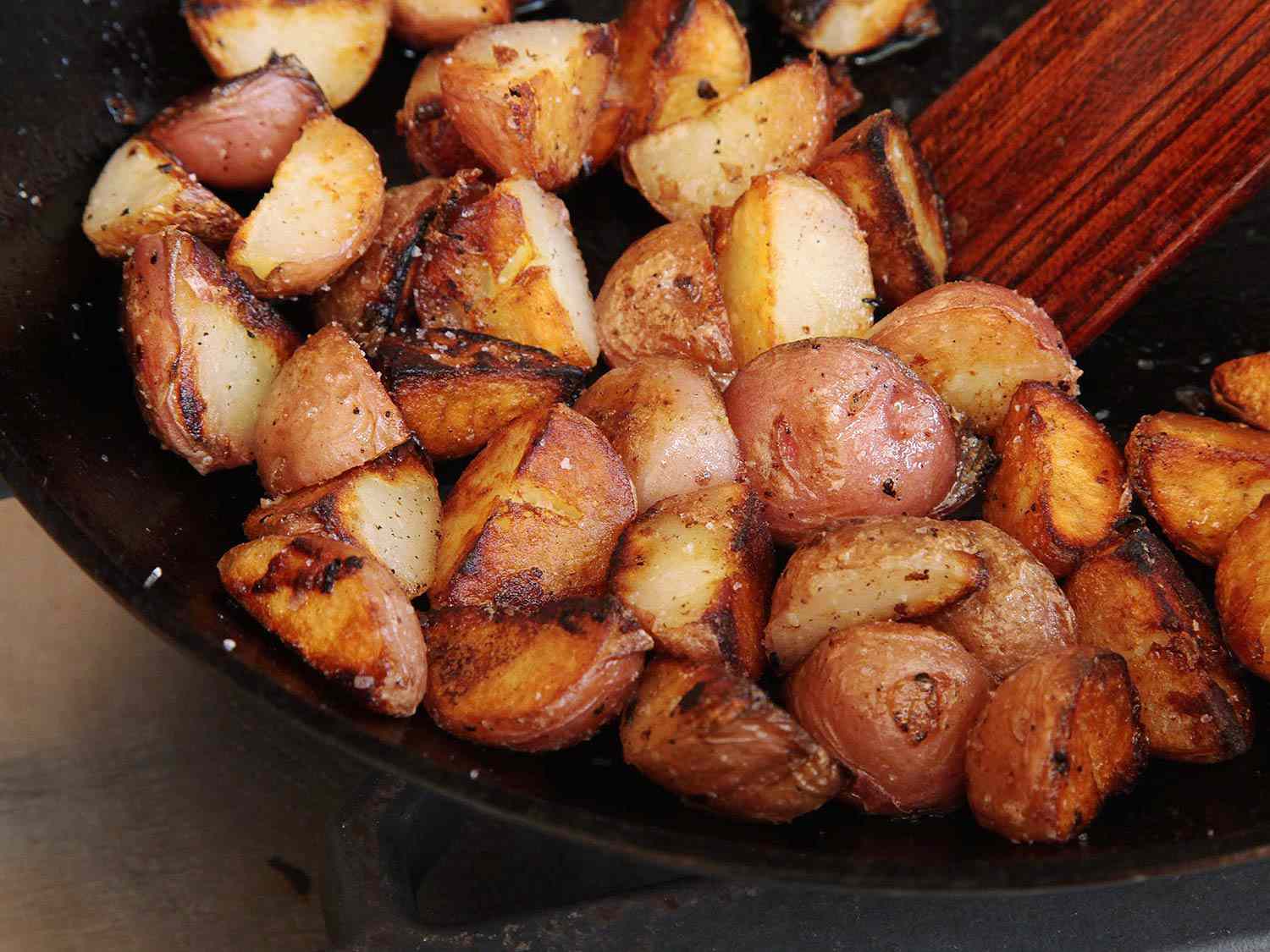 Wooden spoon stirring golden chunks of crispy potatoes in cast iron skillet