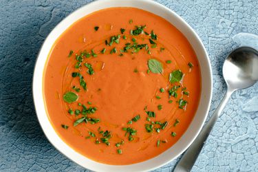 A bowl of gazpacho sprinkled with chopped herbs. The bowl is on a mottled blue surface and there is a metal spoon to the right of the bowl.