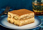 Slice of PSL Tiramisu on a stylish blue and white plate, with a blue tablecloth, sparkling water, and a gold fork