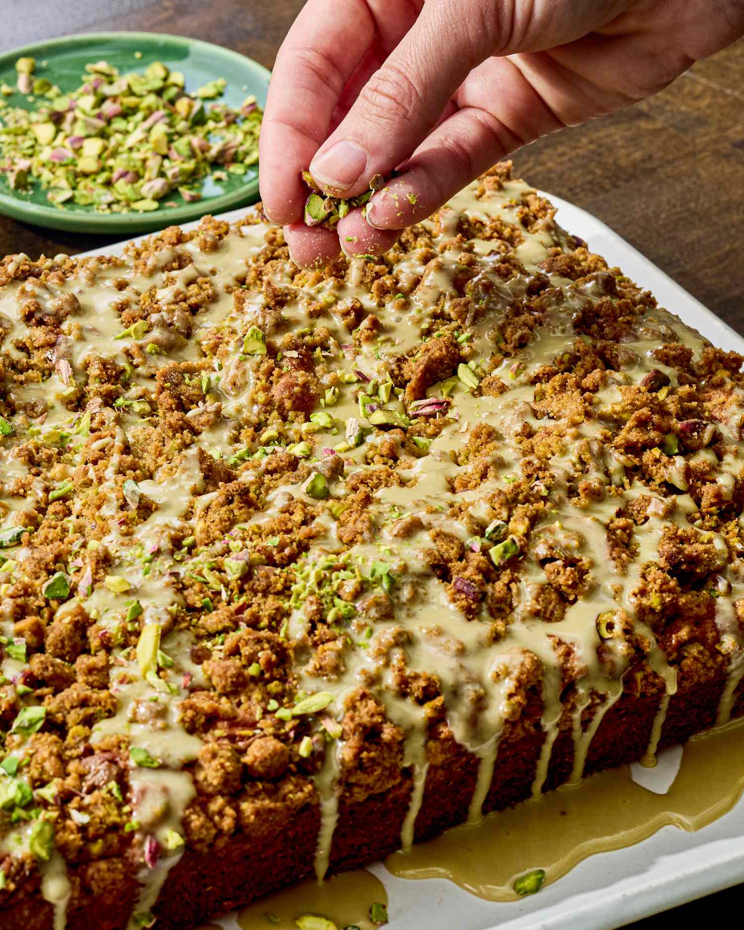 A pistachio coffee cake topped with a drizzle and garnished with pistachio pieces with a hand sprinkling nuts over it