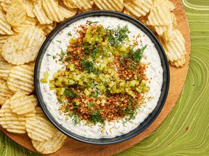 Bowl of pickle dip garnished with panko, dill and pickles, surrounded by a wooden plate full of potato chips, on a green tabeltop