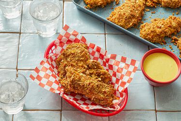 Honey mustard chicken tenders in a basket with dip, more tenders on a tray in the background.