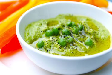 A small white bowl of green pea hummus. On top of the hummus there are a few whole green peas, black pepper and olive oil.