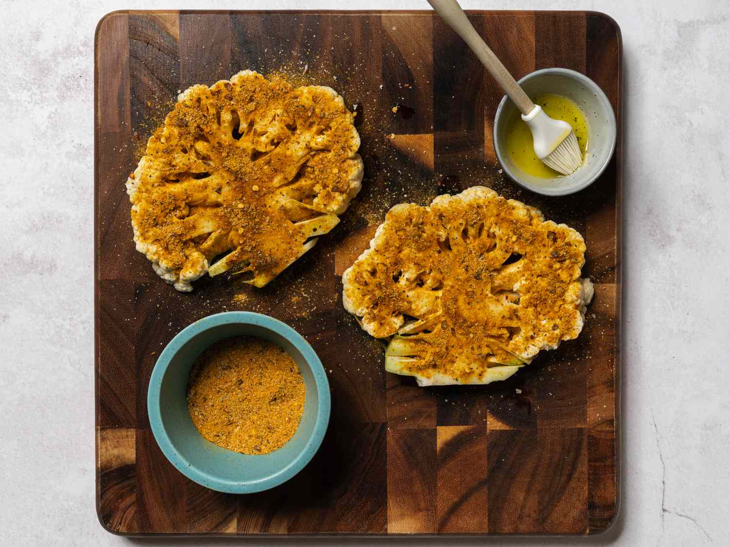 A cauliflower, cut in half lengthwise and sprinkled heavily with the spice mix, on a wood cutting board. The cutting board also has the ceramic bowl holding more spice mix and a small bowl holding olive oil and a silicone brush.